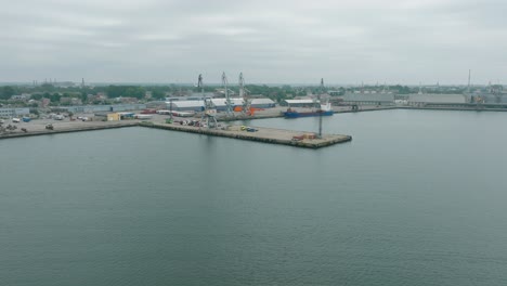 aerial establishing view of port cranes and empty loading docks at port of liepaja , liepaja city in the background, overcast summer day, wide drone shot moving forward, tilt down