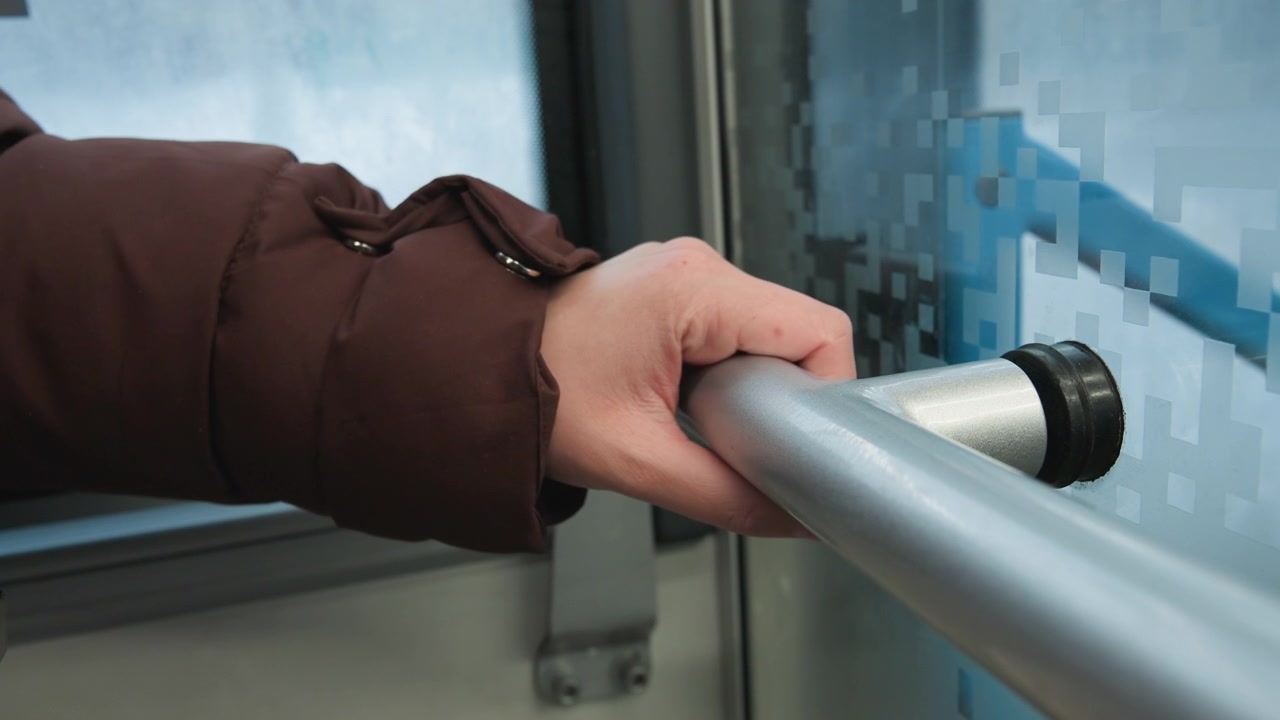 Vista cercana en ángulo bajo de la mano de un adulto mayor agarrando una barandilla horizontal de acero inoxidable, con los dedos curvados y el pulgar presionando la parte superior para mantener el equilibrio dentro de un interior de transporte público luminoso con suaves reflejos