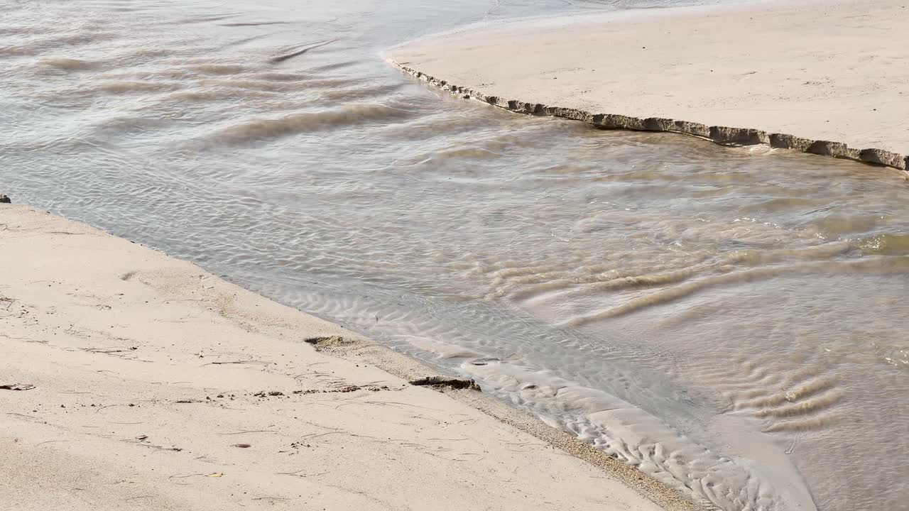 Statisk højvinkelbillede af et lavvandet vandløb, der flyder over sand mod havet.