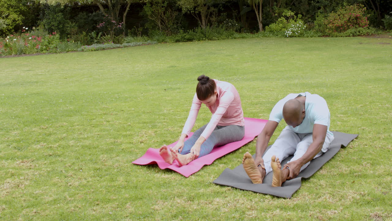 Multiracial couple stretching on yoga mats in park, enjoying outdoor exercise together, copy space