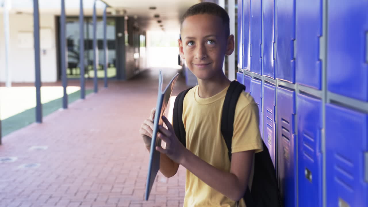 un niño biracial con una mochila está de pie junto a los casilleros azules, sosteniendo una carpeta, en la escuela