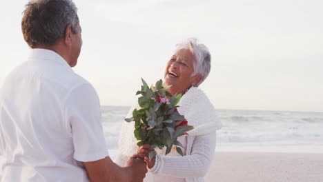 Happy-senior-hispanic-man-proposing-to-senior-woman-with-roses-on-beach-at-sunset