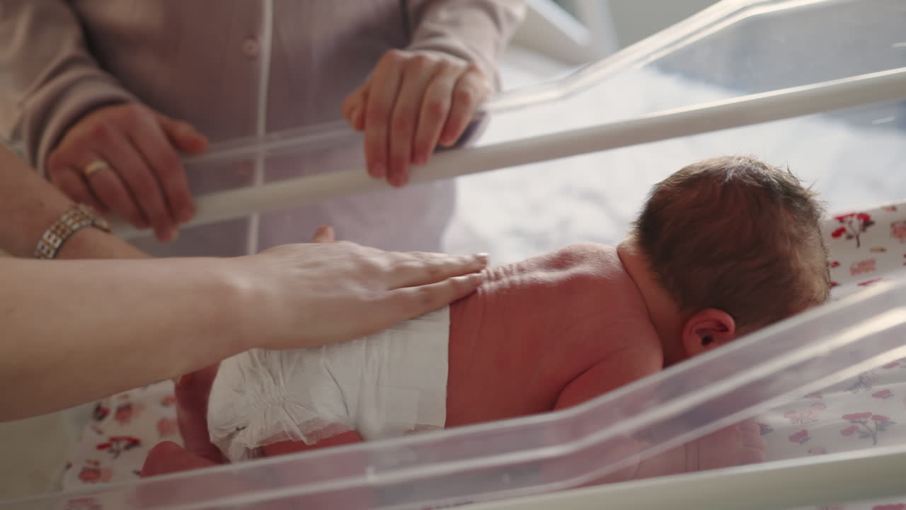 Neonato riposa in una culla di plastica trasparente con le mani dei genitori vicine, a dimostrazione di cura e tenerezza