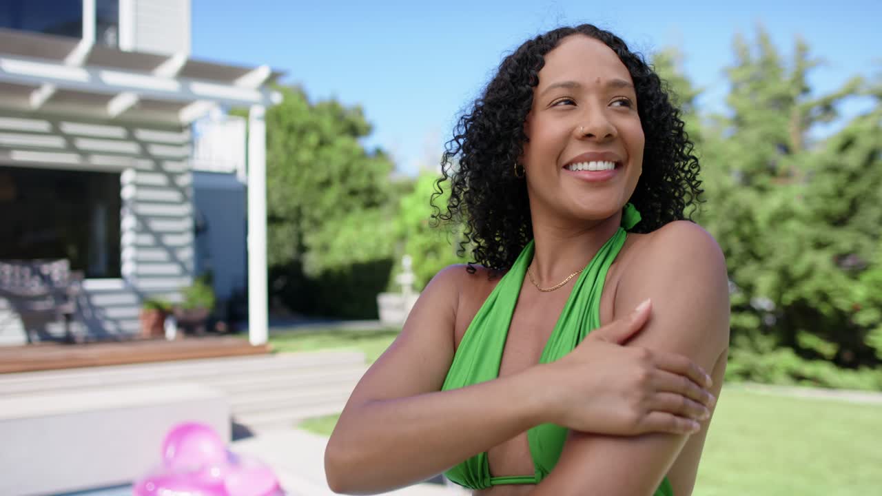 Mulher afro-americana de biquíni verde massageando o ombro e braço direitos no deck da piscina, aquecida pelo sol.