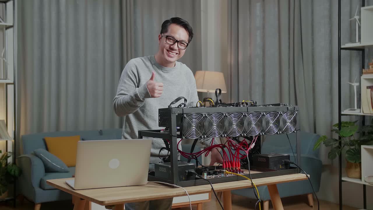 안경을 쓴 미소 짓는 아시아인 남자가 드폰과 채굴 기계를 사용하여 암호화폐를 채굴하는 동안 엄지손가락을 나타니다.