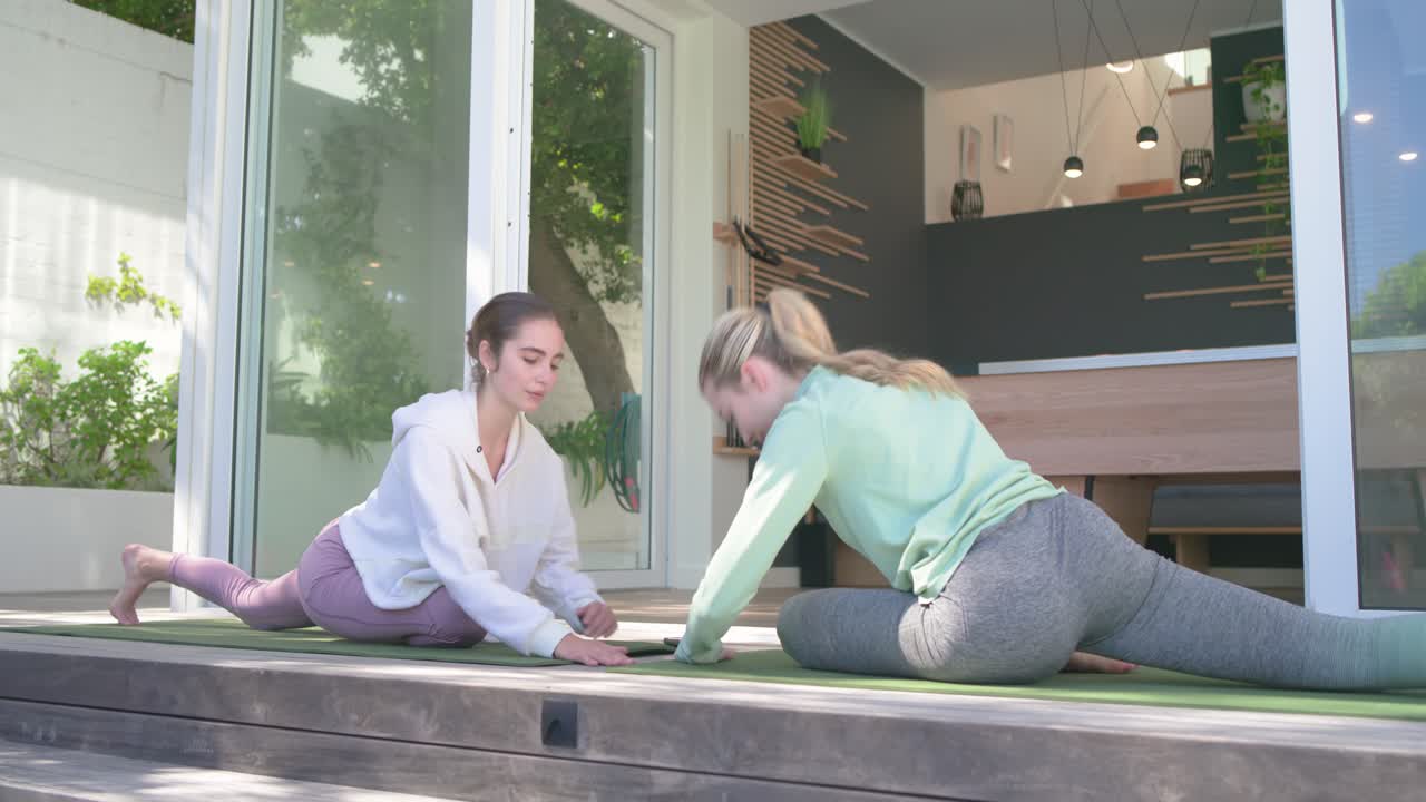 Female friends arching spines on green mats before sliding glass doors then folding forward on deck