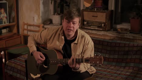 pretty young man playing guitar while sitting on sofa in dark living room