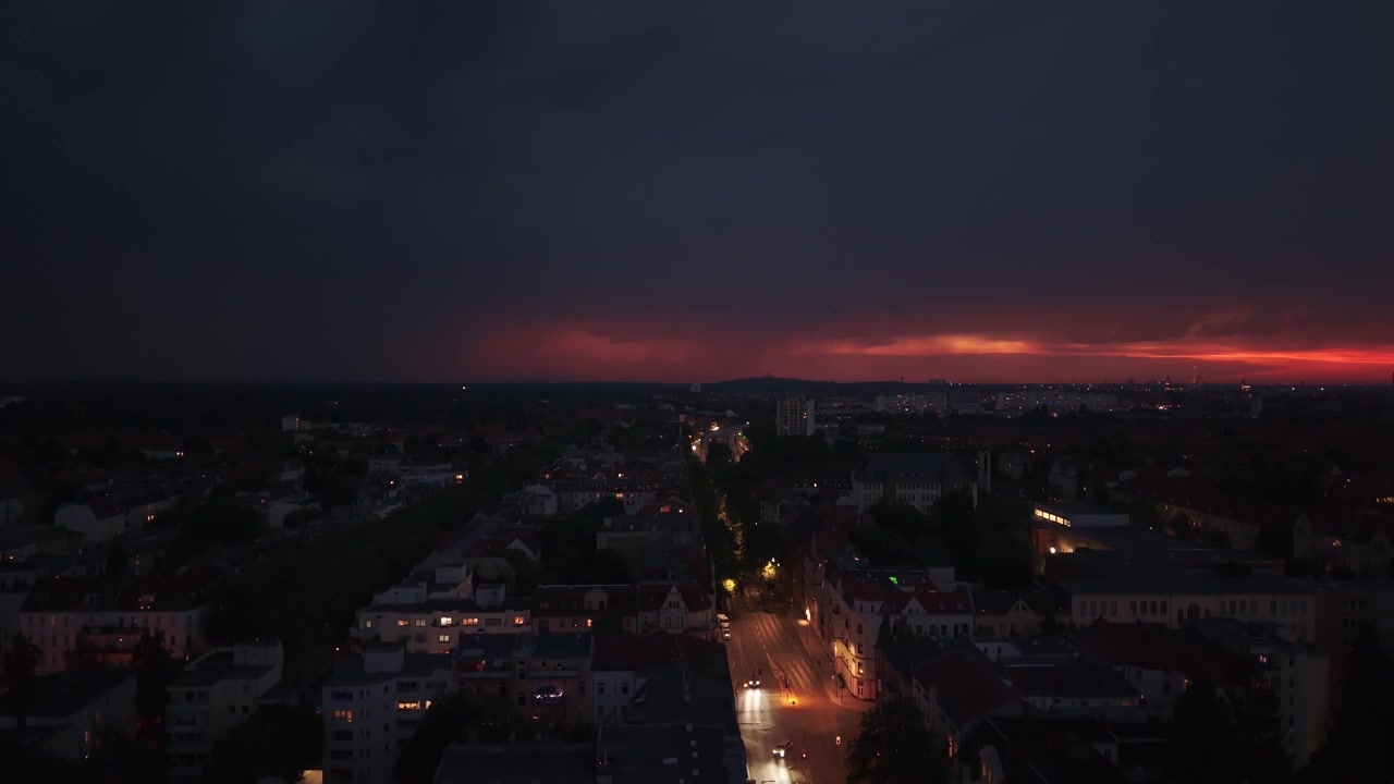 Město s viditelným úderem blesku na bouřkové obloze, osvětlující noční městskou krajinu. Plynulý letecký záběr dronu stoupajícího pro panoramatický pohled.