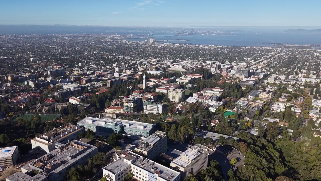 Ampie riprese aeree che mostrano il campus dell'Università della California, Berkeley, con l'iconica Sather Tower e il vasto paesaggio urbano che si estende verso la lontana Baia di San Francisco.