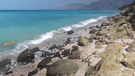 Die-Sonne-Scheint-Auf-Einen-Felsigen-Strand-Mit-Wunderschönen-Steinen,-Der-Bei-Sonnenaufgang-An-Der-Albanischen-Riviera-Von-Türkisfarbenem-Meerwasser-Umspült-Wird