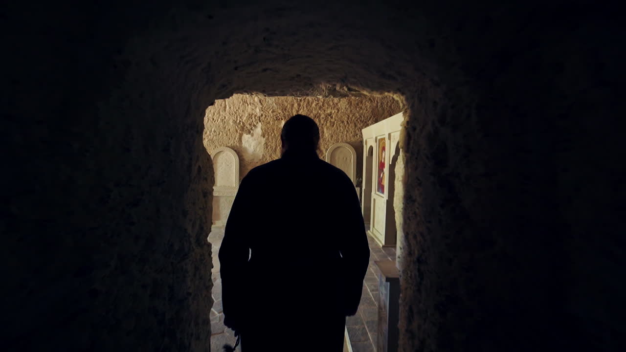 Male silhouette moving by the long corridor with stone walls. Rear view of the monk in black robe entering the chapel in rock monastery.
