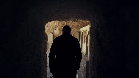 male silhouette moving by the long corridor with stone walls. rear view of the monk in black robe entering the chapel in rock monastery.