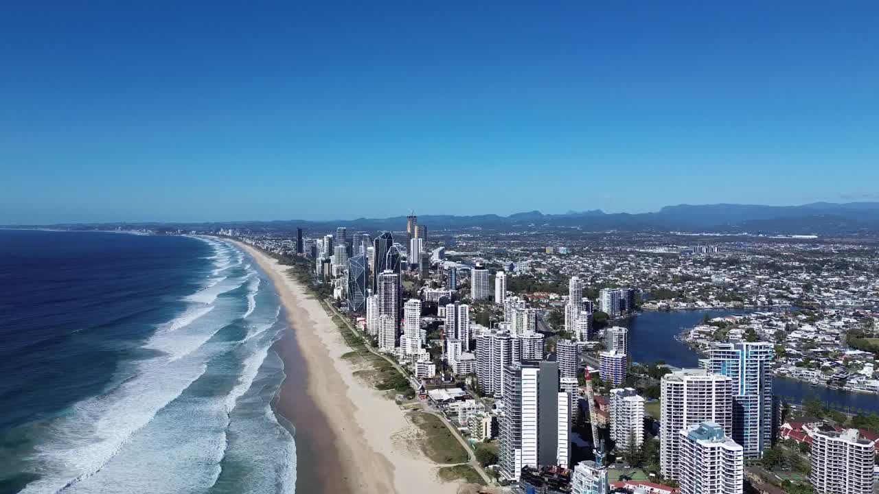 Dramatic drone footage of rough seas crashing against the shore, with towering skyscrapers of the Gold Coast in the background.