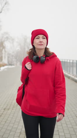 woman with headphones around neck walking along interlocked pathway in red hoodie with bag over shoulder surrounded by snow-dusted park with iron railing benches under foggy winter atmosphere