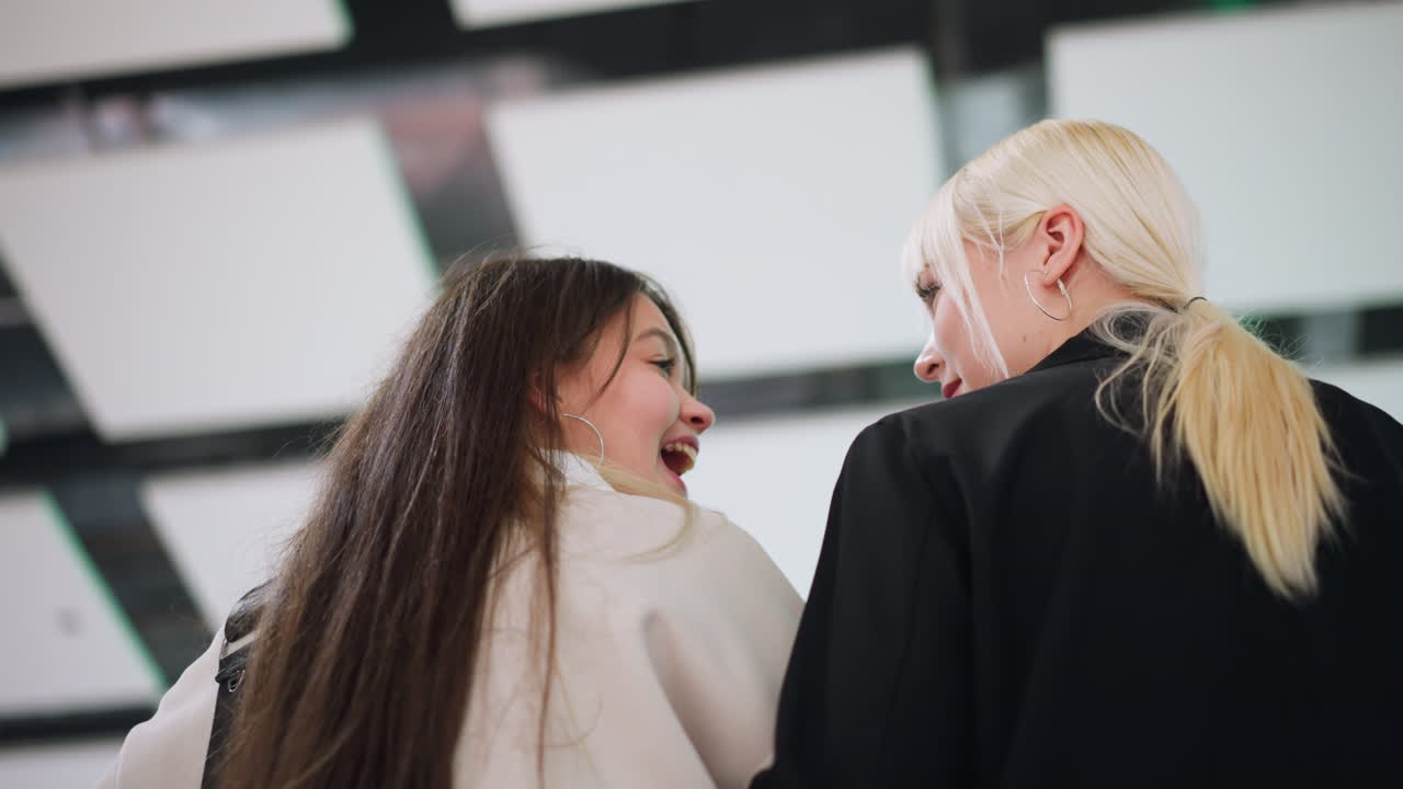 Back view of two women laughing together in casual indoor setting, one with blonde ponytail and other with long brown hair, highlighting friendship, candid joy, and emotional connection