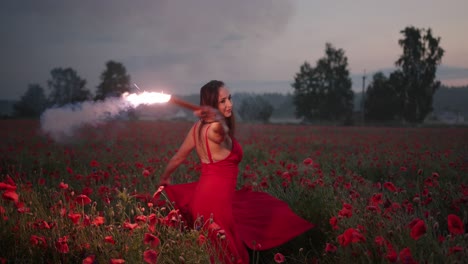 portrait of pretty brunette girl in a red dress dancing with red burning signal flare in poppy field