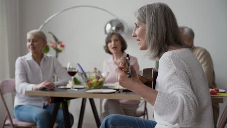 in foreground, happy senior woman singing and playing the guitar sitting on chair, while in blurred background three elderly friends listening to her and singing together sitting at the table