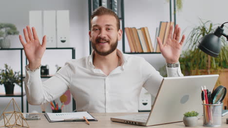 hombre de negocios trabajando en una computadora portátil sonriendo amigablemente a la cámara y agitando las manos saludando en la oficina
