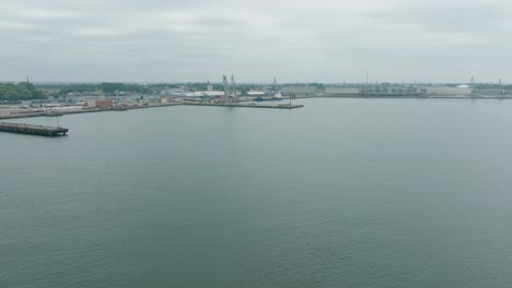 Aerial-establishing-view-of-port-cranes-and-empty-loading-docks-at-Port-Of-Liepaja-,-Liepaja-city-in-the-background,-overcast-summer-day,-wide-drone-shot-moving-forward