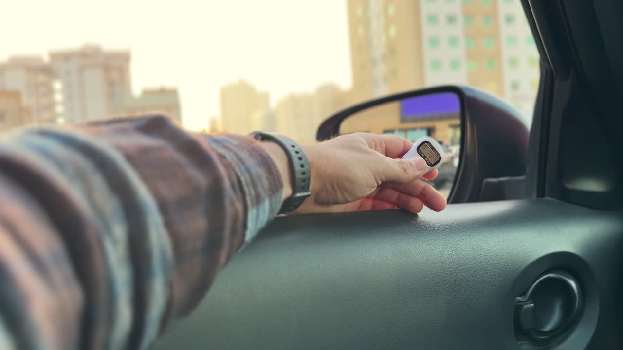 Nærbilde av en sjåførs hånd som holder et lite digitalt telleapparat nær et bilsidespeil med bygninger i bakgrunnen under varmt solnedgangslys, fremhever konseptet transport og sporing