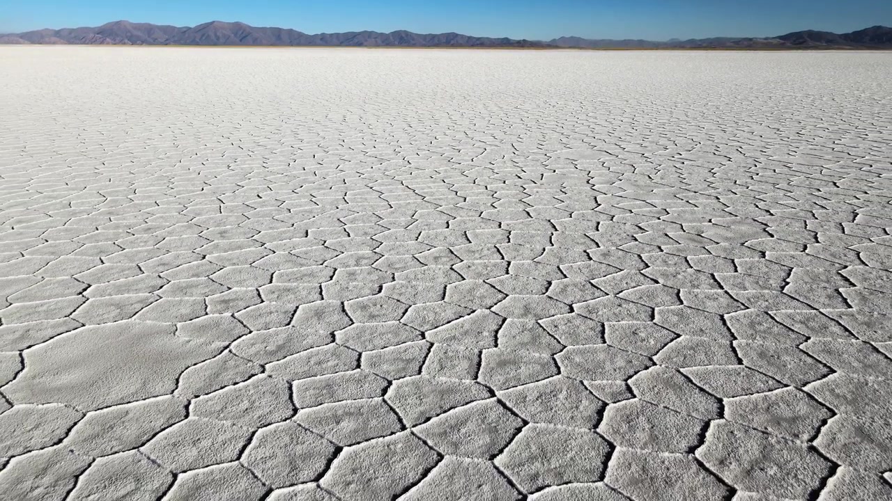 Polygonal cracked salt patterns stretch across Salinas Grandes Salt Flats toward distant mountains under clear blue sky, useful for themes of climate, geology, or lithium extraction.