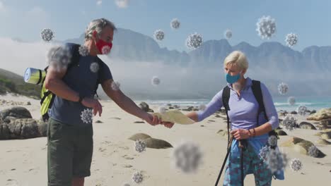 Animation-of-covid-19-cells-floating-over-senior-couple-wearing-face-masks-with-backpacks-on-beach