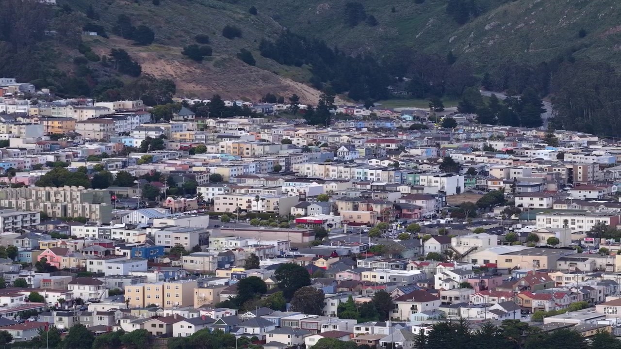 Ripresa da drone che segue, sorvolando le case e gli edifici suburbani fittamente raggruppati del quartiere di Broadmoor a Daly City, California, adagiato contro colline verdi ondulate