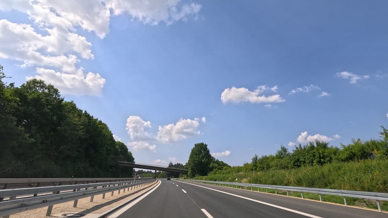 Vehicle travels on empty highway through green countryside under bright blue sky with scattered clouds