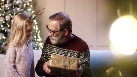 primer plano retrato de un alegre abuelo de pelo gris que recibe un regalo de navidad envuelto de una nieta mientras se sienta en una casa decorada cerca de un árbol de navidad resplandeciente