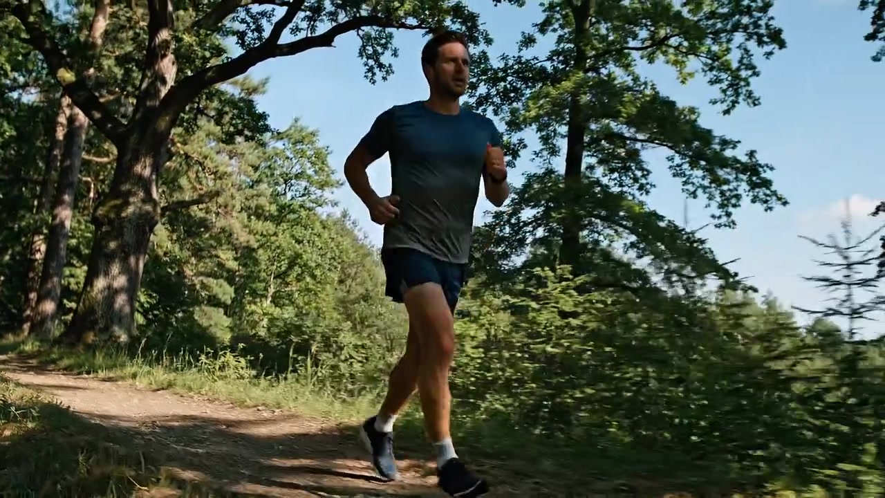Uomo atletico che fa jogging lungo un sentiero forestale soleggiato durante un allenamento fitness all'aperto, circondato da alberi verdi