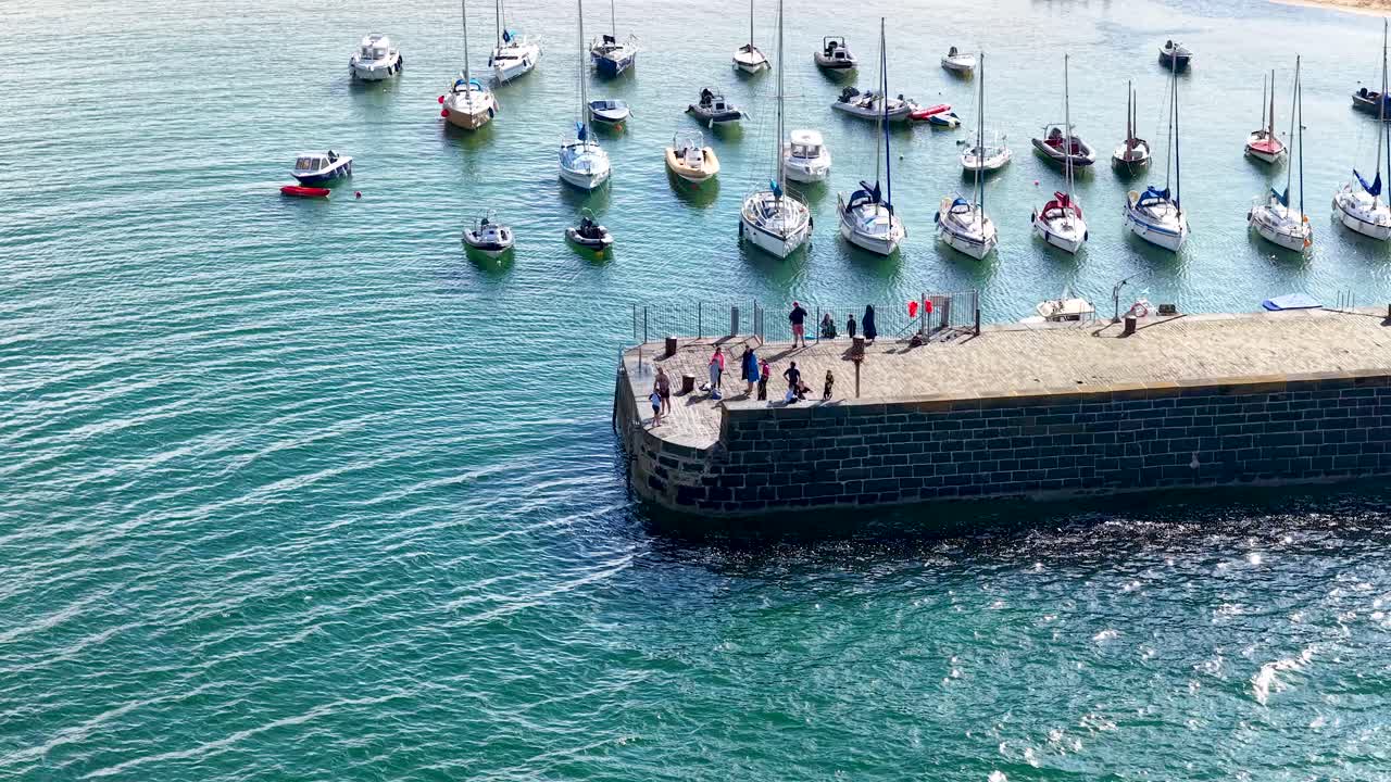 Drone pans over marina with moored sailboats, people gathering on sunlit stone pier, summer daylight