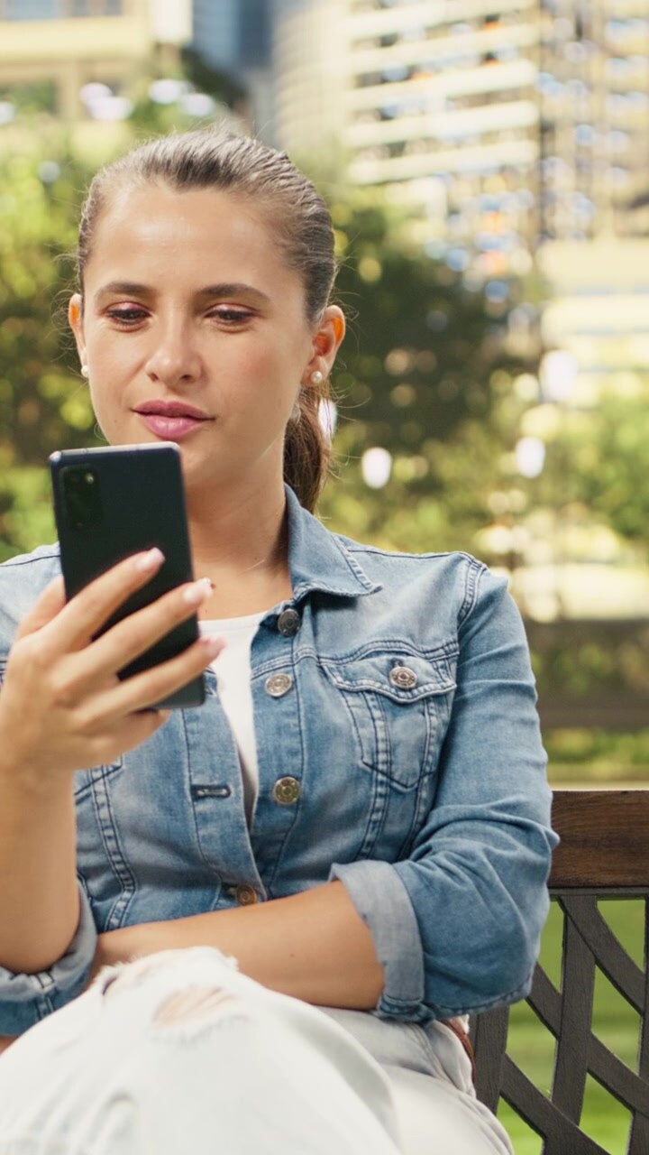 Video verticale. Donna casual che controlla lo smartphone mentre è seduta su una panchina di legno.