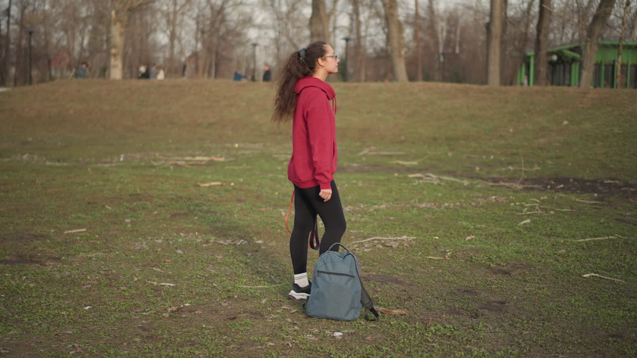 Young Lady Crossing Park Confidently, Young Woman With Backpack And Leash Walking Through Urban Park Confidently, Female Individual Confidently Walking With Backpacks And Leashes Across Park Area