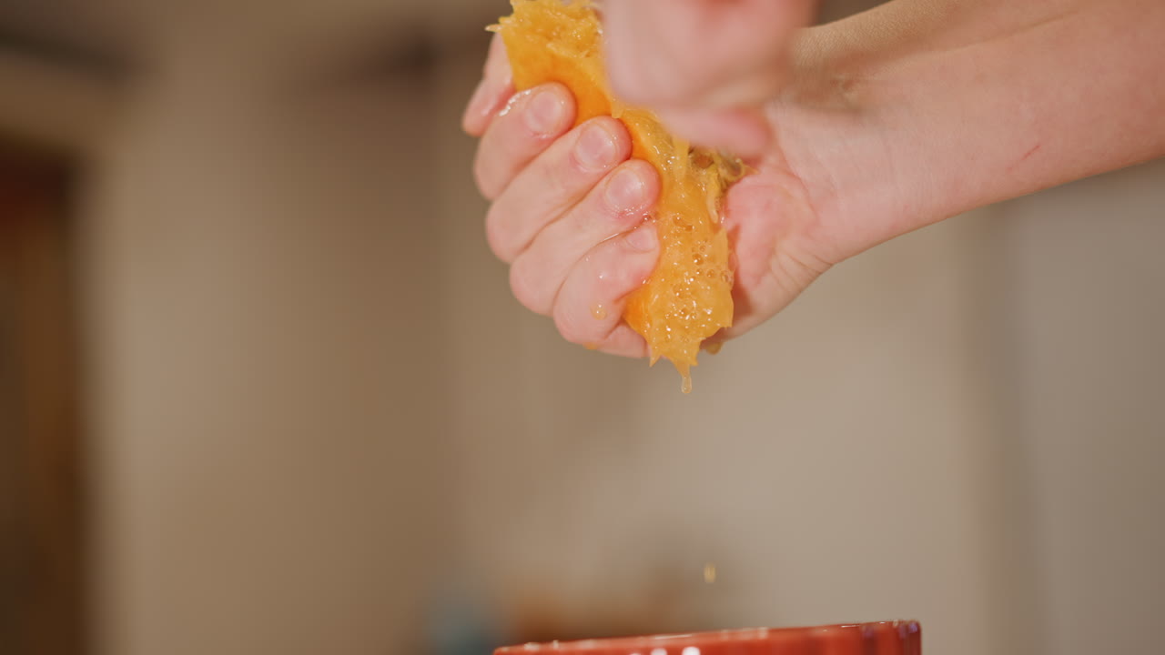 Primo piano di mani che spremono polpa d'arancia. Goccioline d'arancia dense cadono in una tazza di ceramica rossa, bokeh caldo in cucina, consistenza appiccicosa e gocciolamento al rallentatore, preparazione del cibo tattile da parte di un cuoco casalingo, momento sensoriale della bevanda