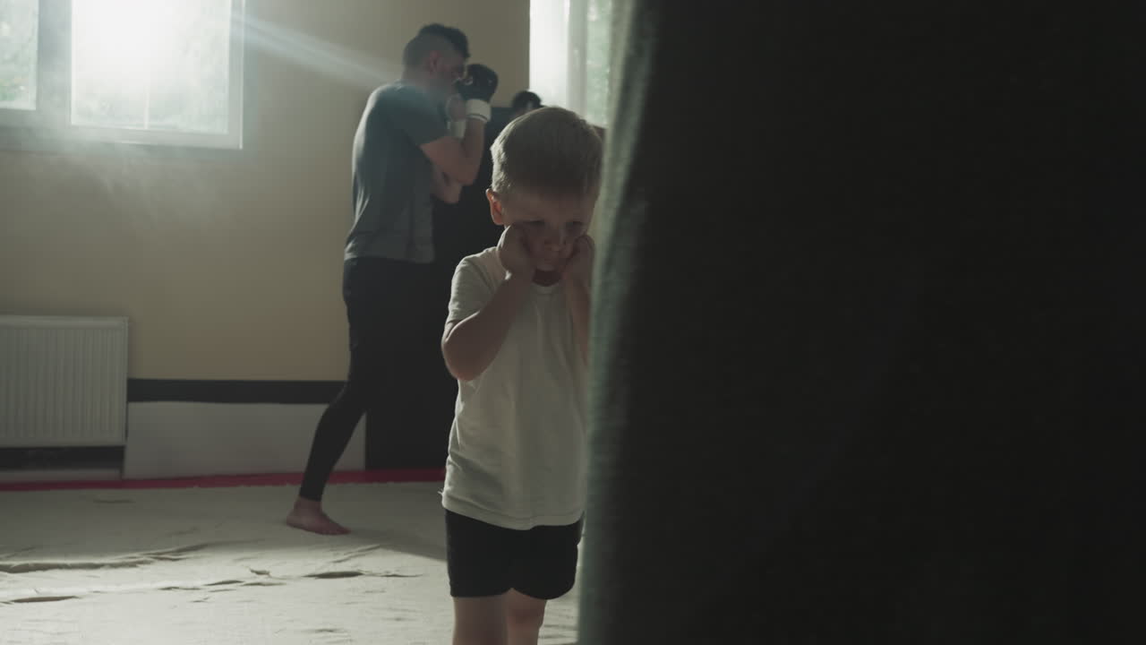 Child kicks punching bag with knee in sports club. Little boy studies kickboxing with adult sportsmen in fighting gym slow motion. Junior athlete