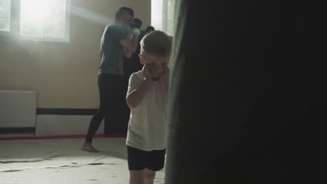 child kicks punching bag with knee in sports club. little boy studies kickboxing with adult sportsmen in fighting gym slow motion. junior athlete