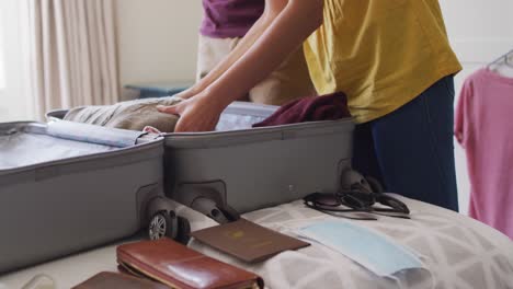 Midsection-of-biracial-couple-packing-clothes-into-suitcase,-preparing-for-travel