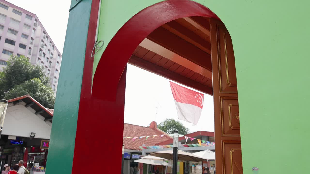 Singapore flag waves behind vibrant archway, urban buildings, daylight, static camera, lively atmosphere