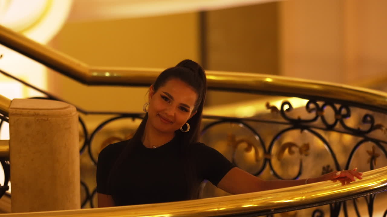 joven y encantadora modelo de dama morena sonriendo mirando a la cámara, junto a la barandilla de la escalera interior de lujo