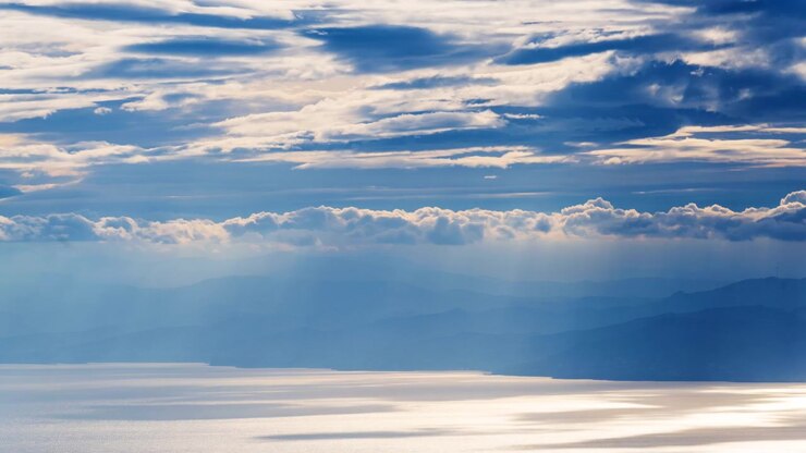 Raggi di sole filtrano attraverso nuvole imponenti su acqua calma e colline distanti all'alba.
