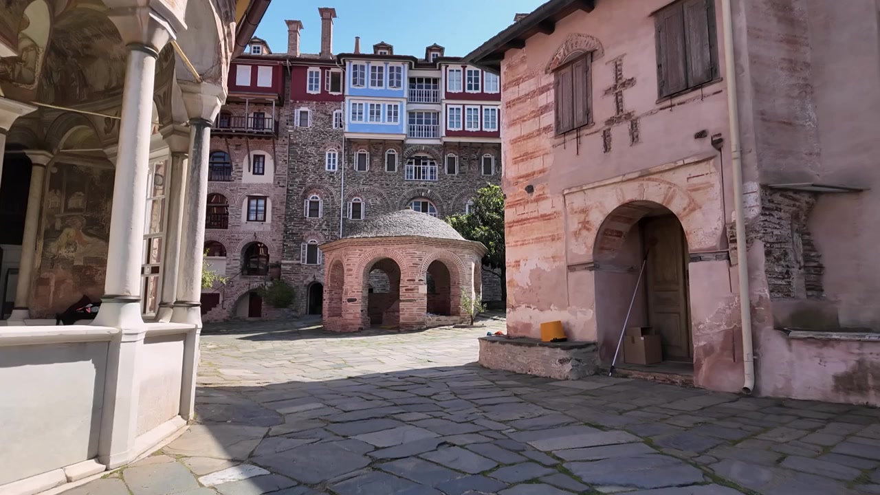 I visitatori passeggiano nel cortile di un antico monastero sul Monte Athos, in Grecia. Il sole illumina gli storici edifici in pietra. I dintorni mostrano un connubio di natura e architettura.