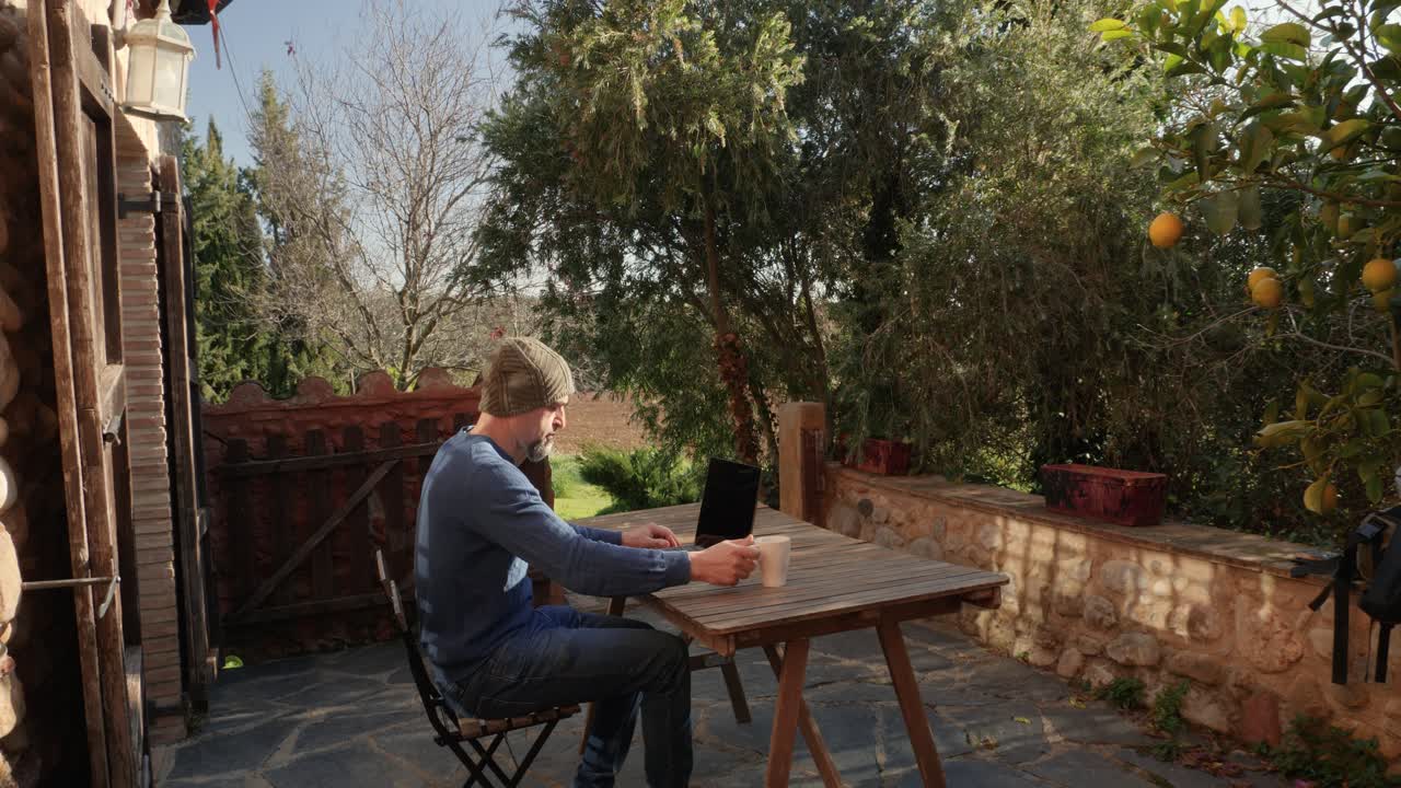 A man sitting at his laptop drinks coffee outside a rural house, with plants sorrounding him