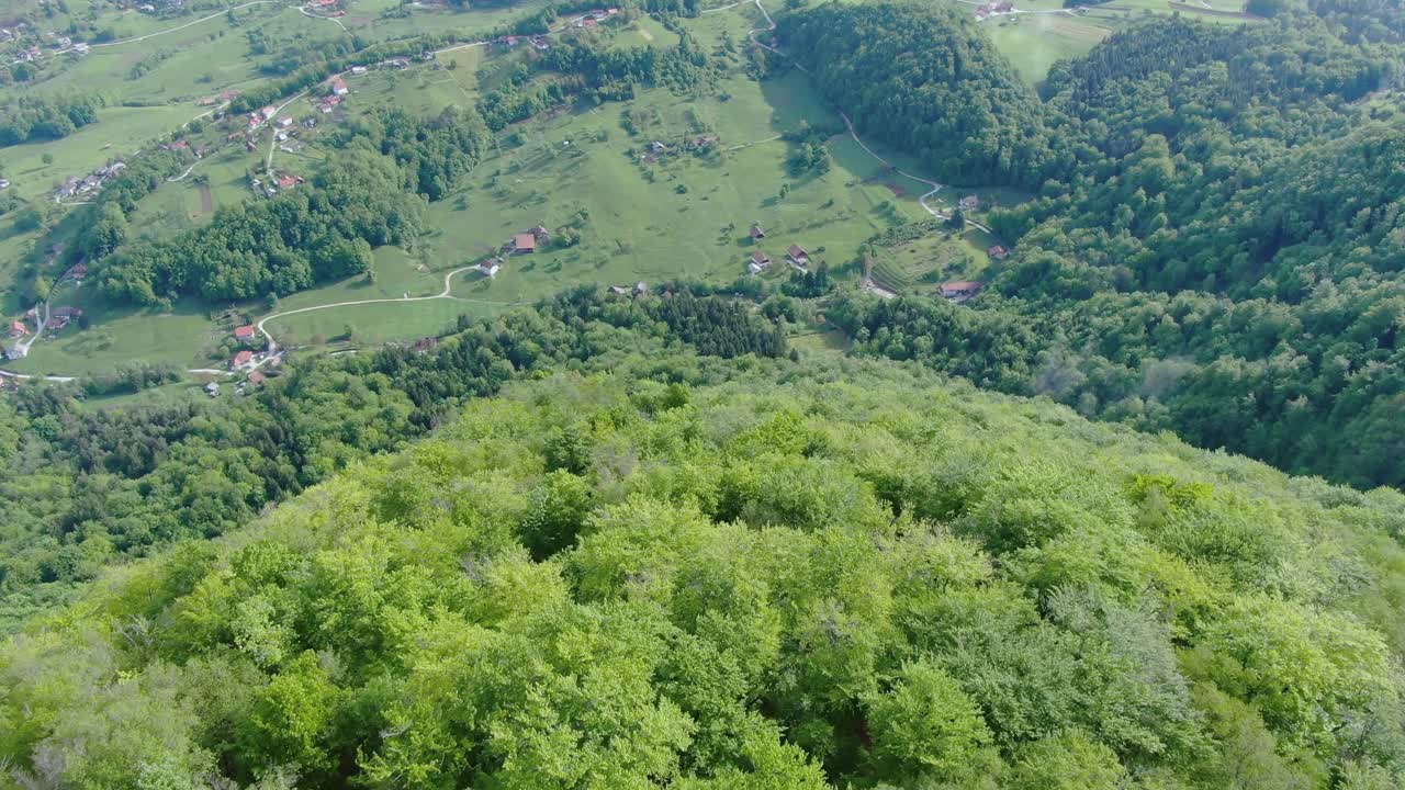 Aerial view up and over tall green trees on hillside to reveal village in valley