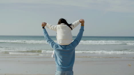 Back-view-of-an-Japanese-father-walking-along-seashore-with-his-daughter-on-his-shoulders,-turning-back-to-the-camera-and-keeping-strolling