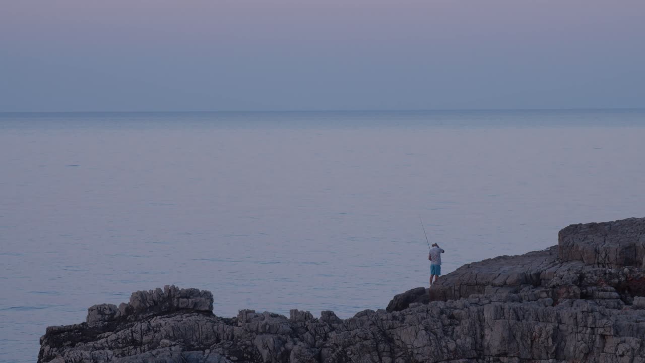 hombre pescando desde una roca junto al hermoso mar, hora azul, toma amplia