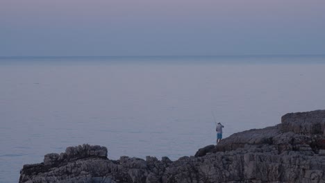 Hombre-Pescando-Desde-Una-Roca-Junto-Al-Hermoso-Mar,-Hora-Azul,-Toma-Amplia