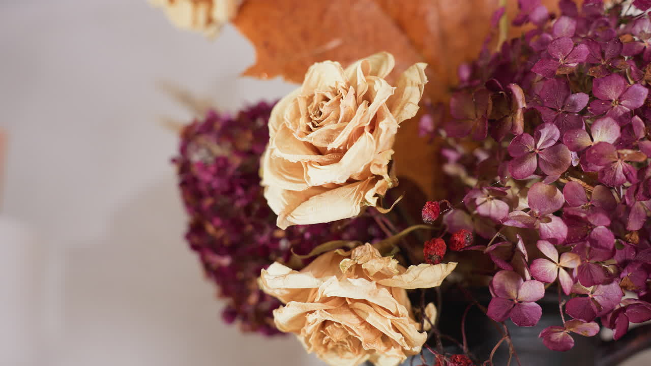 Close up of beige dried roses and purple hydrangeas with autumn leaves and red berries in rustic vase, creating warm natural decor in cozy indoor setting with blurred paper towel roll in background