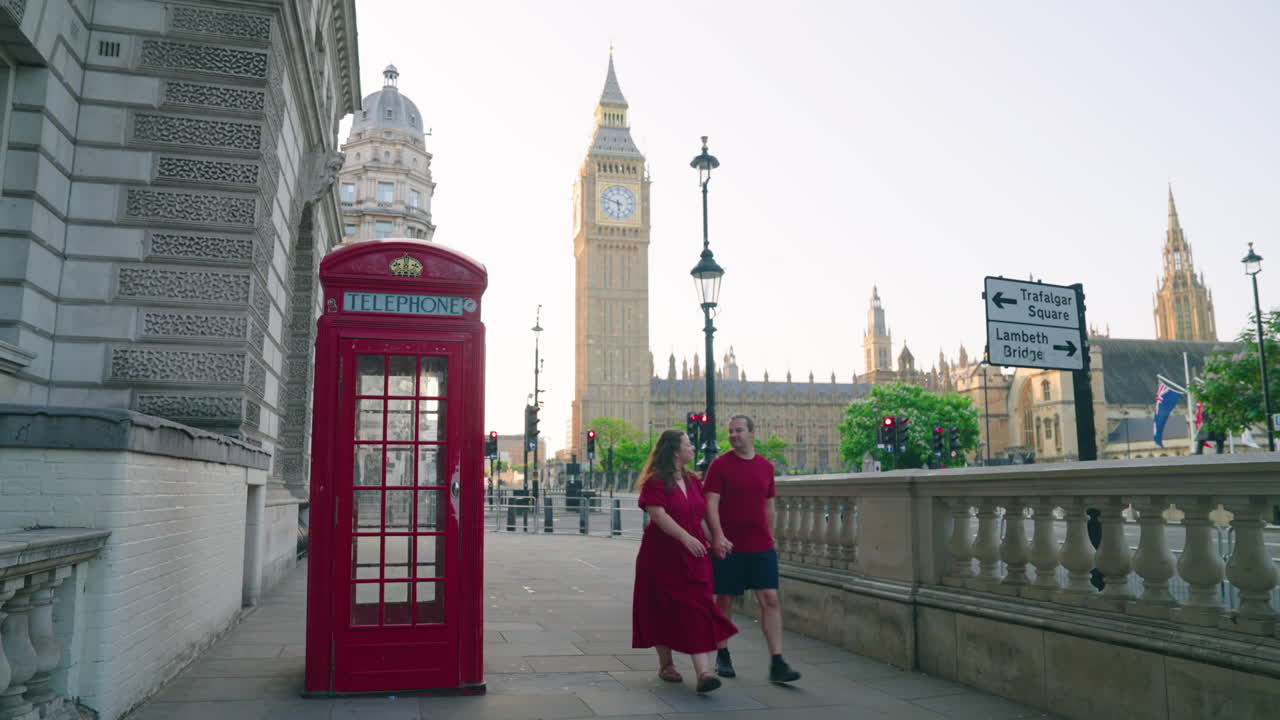 Couple Holding Hands While Walking Next To Red Telephone Box With Big Ben In The Background In London, UK. - wide shot