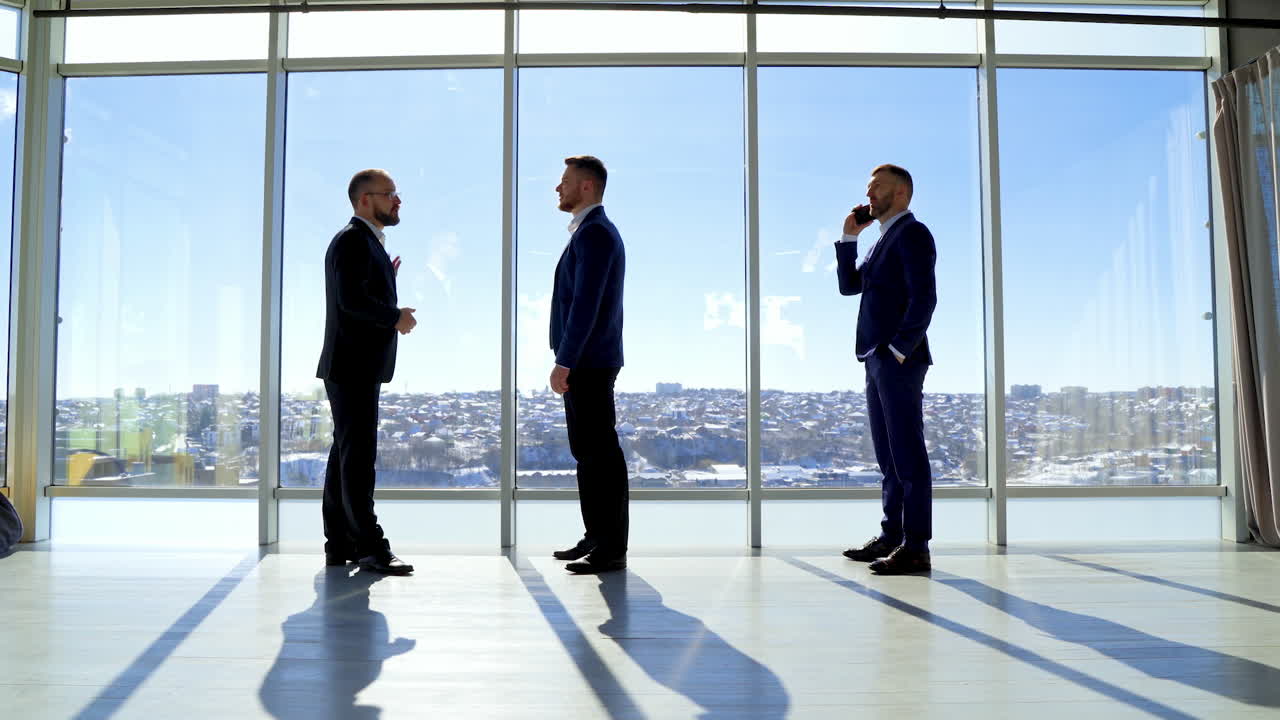 Full-length portrait of business people. Male entrepreneurs in elegant suits standing in modern office and talk about business. Profile view. Panoramic windows view on city.