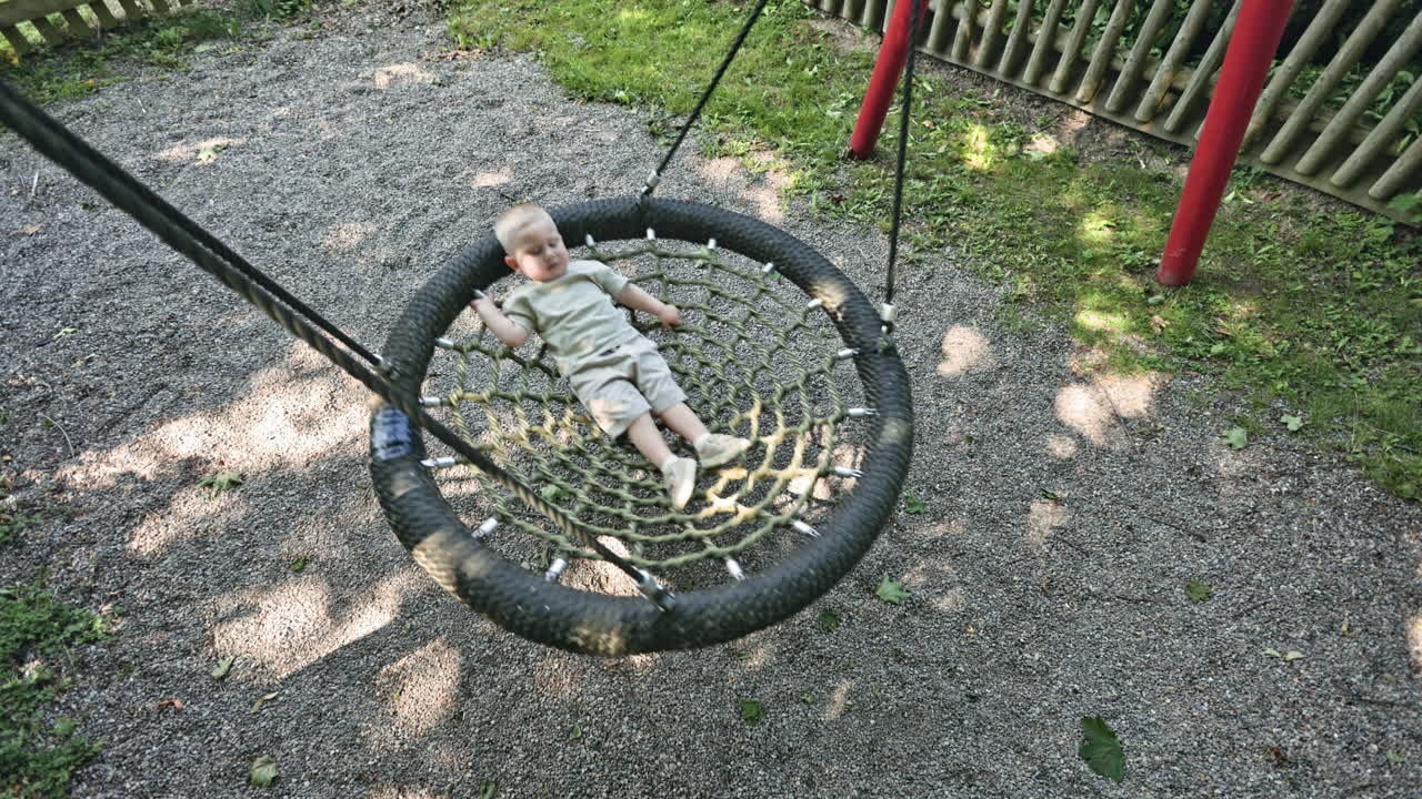 A 2 year old toddler in shorts swings on a spider web swing on a warm summer day. A toddler boy swings on a playground in a place. Entertainment for a child on a playground